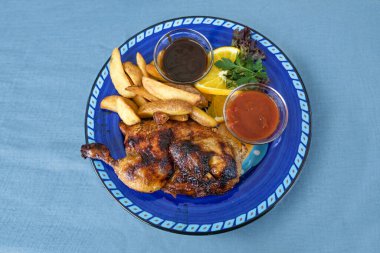 Izgara yarım tavuk, patates dilimleri ve iki soslu rustik mavi tabak ve masa örtüsü, fotokopi alanı, yukarıdan yüksek açılı görüş, seçilmiş odaklanma.