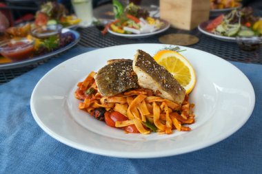 Kırmızı pesto, domates ve zeytinli kavrulmuş morina balığı, portakal, limon ve dereotu garnitür, beyaz tabak ve mavi peçete bir masada arkadaşlarla bir akşam yemeğinde, seçilmiş odak, dar bir alan derinliği