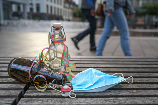 Party waste in the city during covid 19 restrictions, liquor and beer bottles with a surgical face mask carelessly left on a wooden bench in the busy market place, while people passing by, copy space, selected focus, narrow depth of field