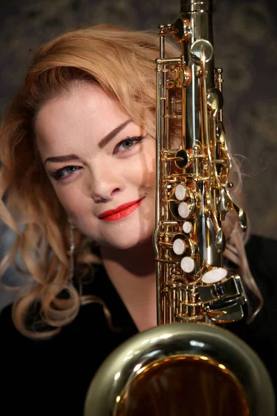 Portrait of a charming blonde with a saxophone in her hands. Beautiful girl in a black evening dress. Young woman with her hair and bright red lips. Face close up. Saxophonist lady