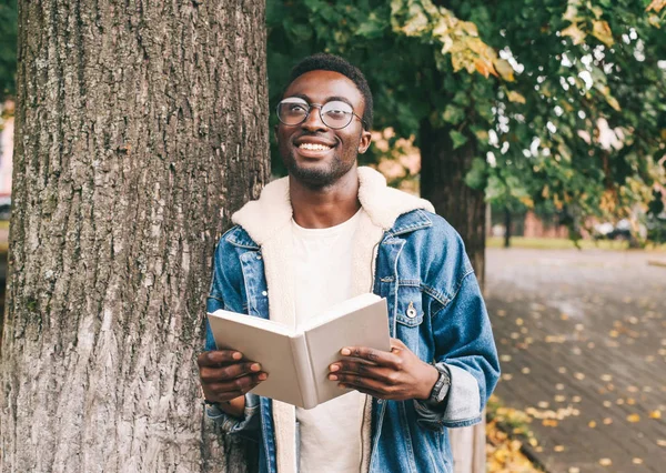 Happy smiling african man dreams, holds book in autumn city park near tree