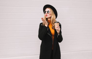 Güzel kadın üfleme kırmızı dudaklar tatlı hava gönderme üzerinde siyah ceket palto, yuvarlak şapka, giysi gri duvar arka plan kahve fincanı ile öpmek