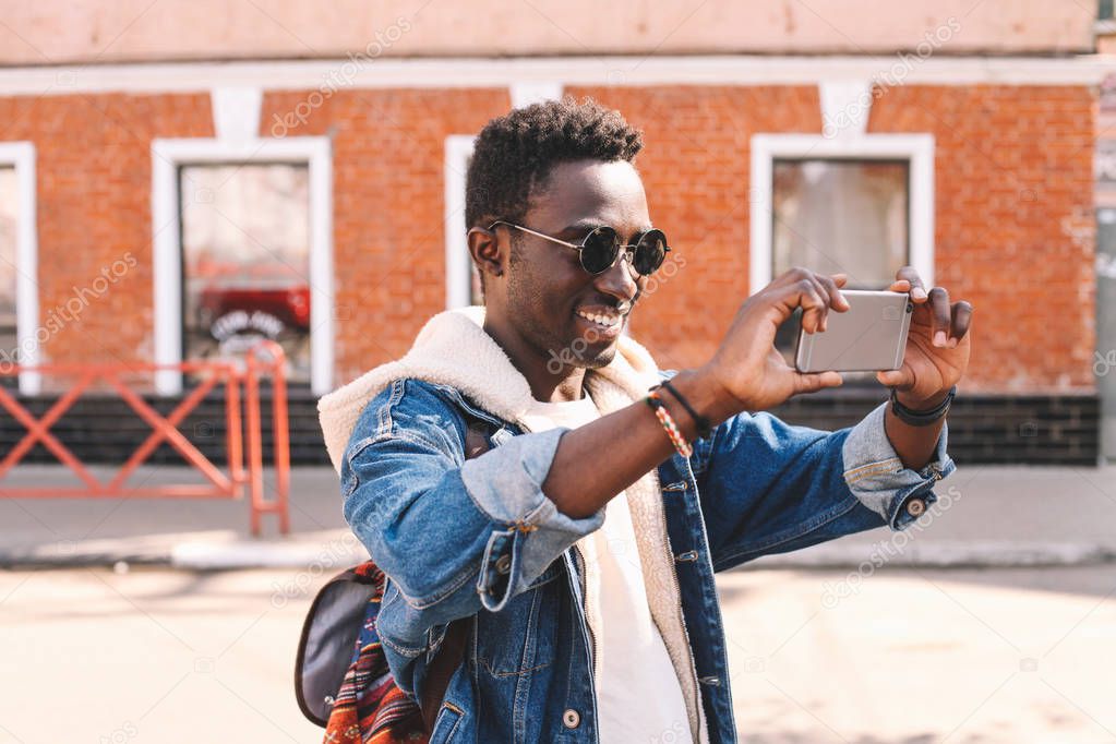 Cheerful smiling african man taking selfie picture by smartphone walking on city street