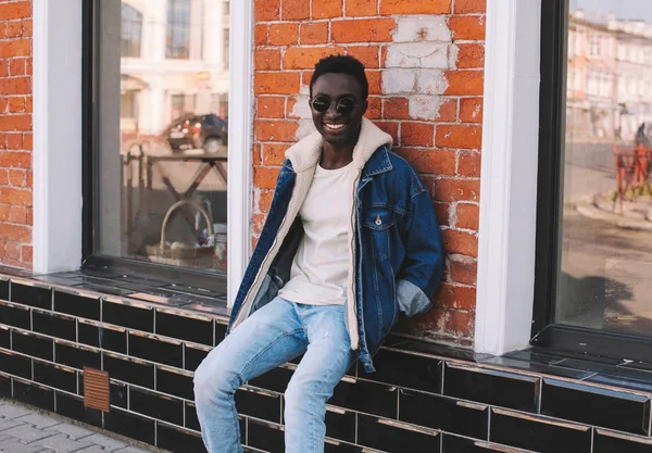 Stijlvolle gelukkig glimlachend Afrikaanse man dragen jeans jasje zitten o — Stockfoto