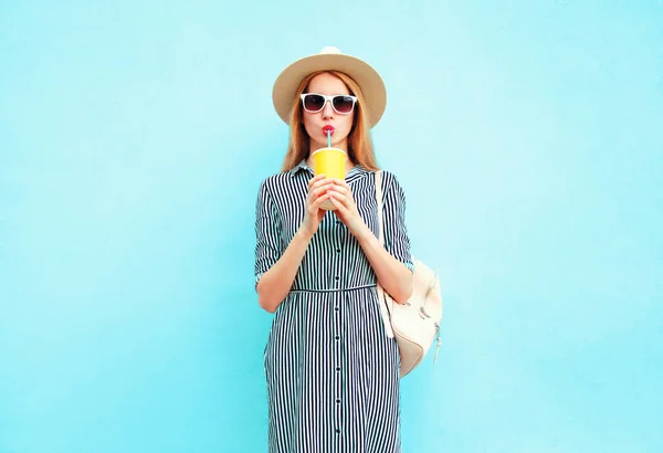 Mladá žena pije ovocnou šťávu z kelímku, nosí v létě kulatý slaměný klobouk, pruhovaný oděv na pozadí modré zdi — Stock fotografie
