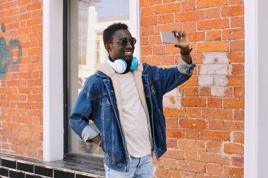 Mutlu, havalı, gülümseyen Afrikalı adam, duvarın arkasında, akıllı telefondan selfie çekiyor.