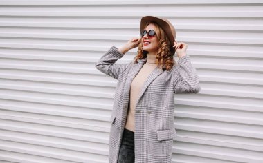 Gri ceketli, mutlu, şık bir kadın, yuvarlak şapkalı, beyaz duvardaki boş fotokopi odasına bakıyor.