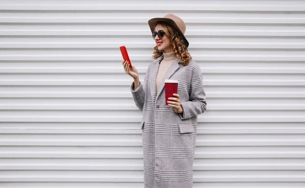 Mulher sorridente feliz elegante segurando xícara de café olhando para o telefone vestindo casaco cinza, chapéu redondo no fundo da parede branca — Fotografia de Stock