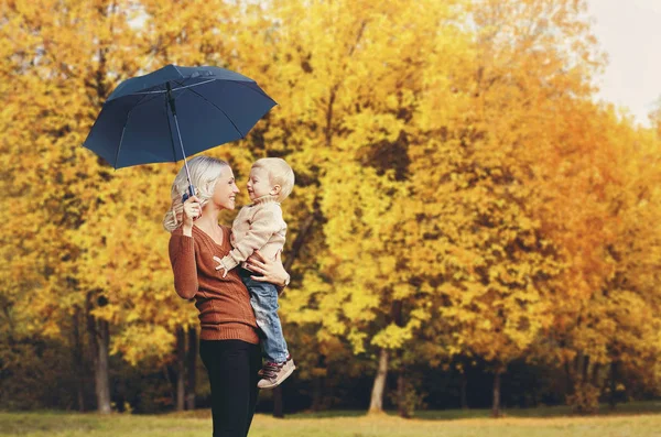 Feliz sorrindo mãe segurando criança com guarda-chuva se divertindo juntos ao longo de árvores de outono fundo — Fotografia de Stock