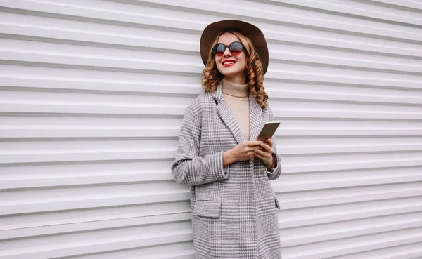 Retrato elegante feliz sorrindo mulher segurando telefone vestindo casaco cinza, chapéu redondo olhando para longe sobre fundo da parede branca — Fotografia de Stock