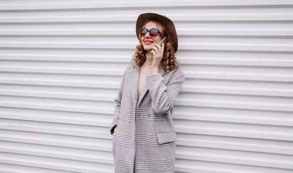 Retrato elegante mujer sonriente llamando en el teléfono inteligente con abrigo gris, sombrero redondo mirando hacia otro lado sobre fondo blanco de la pared — Foto de Stock