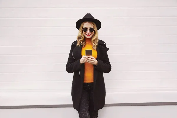 Stylowa uśmiechnięta kobieta patrząca na telefon w czarnym płaszczu, okrągłym kapeluszu, okularach przeciwsłonecznych w mieście na szarym tle ściany — Zdjęcie stockowe