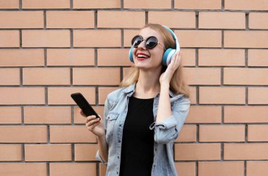 Telefonlu, kablosuz kulaklıklı, siyah yuvarlak şapkalı, kot ceketli, duvar arkasında müzik dinleyen mutlu bir kadının portresi.