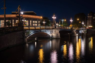 Amstel nehri üzerinde bir köprü ve ışıklar gece vakti kanal suyuna yansıyor, Amsterdam, Hollanda.