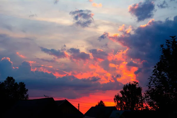 White clouds in the light blue sky and dark blue clouds in red and orange sky looking like fire over black silhouettes of roofs of country houses and trees on summer evening