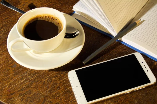 Bir fincan kahve ve çay kaşığı tabağın üstünde defter, kalem ve ahşap bir masanın kahverengi yüzeyinde akıllı telefon.