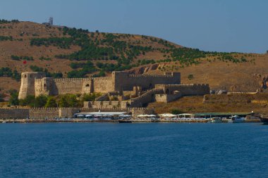 Türkiye 'nin Bozcaada (Tenedos) kıyılarının manzarası. Resimde bir ortaçağ kalesi, turist dükkanları, sahil şemsiyeleri ve kıyı boyunca bazı tekneler yer alıyor..
