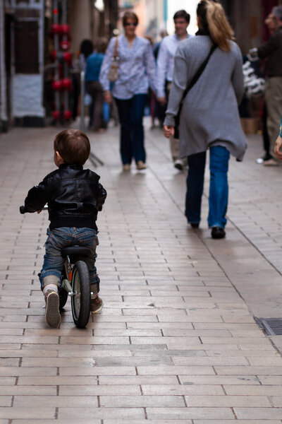 Close up image showing a cute little boy wearing cool stylish jeans and a leather jacket, riding a bike in a busy street. He is following his mom at a distance and is comfortable being in public .