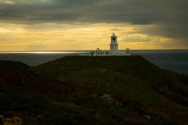 Atlantik okyanusuna bakan bir uçurumun tepesindeki izole bir deniz fenerinin görüntüsü. Gün batımında Galler 'deki St. Anne' s Head Pelerini 'nden çekilmiş. Engebeli arazi ve binaya giden küçük bir yol görülüyor.