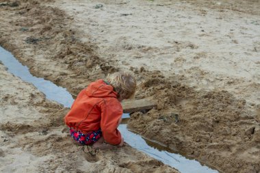 Küçük tatlı sarışın bir kız kumların üzerinde oynuyor. Deniz suyunun aktığı küçük bir su yolunda çömeliyor. Kumu kazıyor ve ilginç bir şekilde yüzeyin altında ne olduğunu araştırıyor.