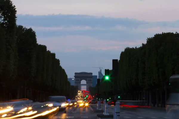 A long exposure low light image of the famous Champs-Elysees avenue in Paris with the iconic Arch in the background. Trees are seen as silhouettes on each side. Beautiful sky is red with clouds.