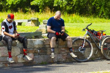 Maryland, ABD 06 / 27 / 2020: İki adam ormanda taş bir duvarda dinleniyor ve Eachoter 'la konuşuyor. Bisiklet kıyafetleri giyiyorlar ve zorlu bir yolu daha yeni bitirdiler. Çamura bulanmışlar. Bisikletlerinden biri duvarın yanında park halinde görülmüş..