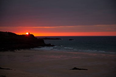 St. Ives yakınlarındaki Cornwall 'ın engebeli sahilinden çekilmiş Atlantik Okyanusu üzerinde bir günbatımı görüntüsü. Fotoğrafta bir körfez, kumlu sahil, sakin deniz, bulutlu gökyüzü, kaya siluetleri ve ufuk görünüyor..