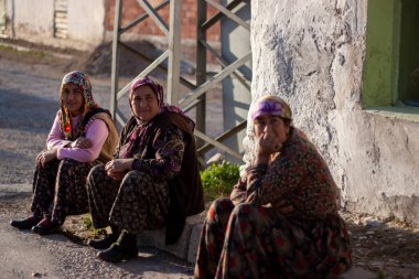 Manisa, Türkiye: Bir tarım köyünde yaşayan üç Türk kadın eski bir evin kaldırımında oturuyor. Geleneksel boyalı Türk elbiseleri ve başörtüleri giyiyorlar.