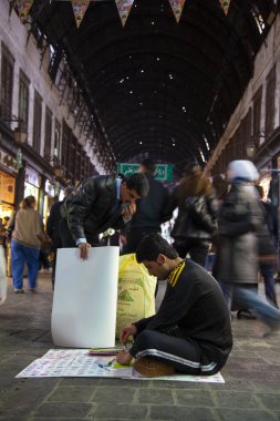 Şam, Suriye 03 / 28 / 2010: Genç bir Suriyeli, geçimini sağlamak için vitrininde ucuz plastik çizim araçları satmaya çalışan kalabalık El-Hamidiyah Souq 'un zemininde bacak bacak üstüne atmış bir şekilde oturuyor.