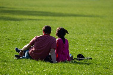 Genç bir çift, güneşli bir öğleden sonra çimlerin üzerinde oturup parkta dinlenirken biraz boş zaman geçiriyor. Erkek tişört ve kot pantolon giyiyor, kadın pembe bluz giyiyor ve botlarını çıkartıyor..