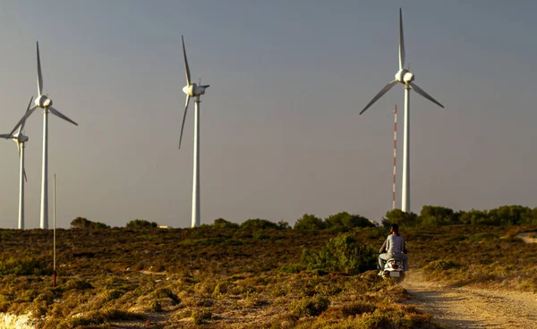 View of a young couple riding a scooter motor bile on a dirt road towards the wind turbines  through maquis shrubland. Image shows the wind farm iof Bozcaada at sunset in the background.
