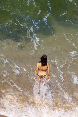 Bikini giyen güzel, uzun saçlı, beyaz bir kızın hava görüntüsü sahilden denize doğru yürüyor. Sıcak bir yaz günü. Suyun içinde yürürken etrafında su sıçrıyor..