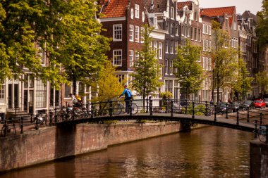 Amsterdam, Hollanda 05 / 15 / 2010: Amsterdam 'da bir kanal üzerindeki yaya köprüsünün görüntüsü. İki bisikletçi bisikletleriyle oradan geçiyorlar. Kanalın yanında geleneksel Hollanda dar evleri var..