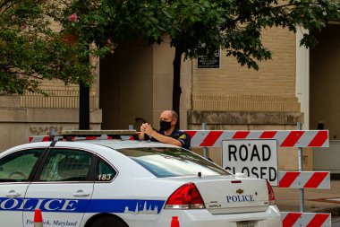 Frederick, MD, ABD 08 / 14 / 2020: Görevdeyken genç bir beyaz erkek polis memuru maske takıyor. Polis arabasının yanında yol bariyerinin önünde duruyor..