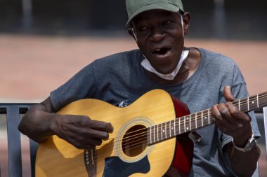Annapolis, MD 08 / 21 / 2020: Beyzbol şapkası ve maske takan yaşlı bir Afro-Amerikan adam klasik gitar çalıyor ve sokakta bir bankta otururken şarkı söylüyor. O bir sokak müzisyeni..