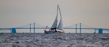 Arka planda ünlü Bay Köprüsü 'nün siluetiyle Chesapeake Körfezi' nden geçen bir yelkenlinin panoramik görüntüsü. Teknede gün batımının tadını çıkaran insanlar var.,.