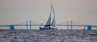 Panoramic image of a sailboat moving across Chesapeake Bay with the silhouette of the famous Bay Bridge in the background. There are people on the boat who are enjoying the sunset over the bay,. clipart