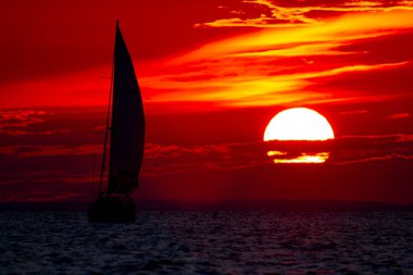 A spectacular sunset over Chesapeake Bay Maryland as captured from the east shore. Image features the large sun going down among clouds on a red sky and the silhouette of a sailboat with full sails. clipart