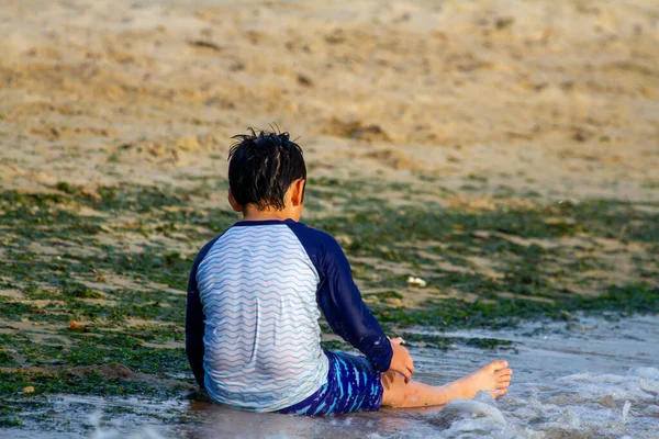 A young caucasian boy wearing rash guard long sleeve swimsuit is sitting on sand by the sea. He is playing alone with the sand and water. Image was taken in Rockhall, coast of Chesapeake bay, maryland