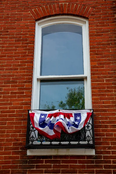 A narrow vertical arched window with wooden frame located on a vintage brick building. The window has metal railings on the lower half which is decorated with a cotton bunting  with American Flag colors on it.