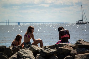 Rock Hall, MD, ABD 08 / 30 / 2020: Güneşli bir yaz gününde üç genç kız (bir Afrikalı Amerikalı ve iki Kafkasyalı), Chesapeake Körfezi 'nin yanındaki kayalıklarda oturuyorlar. 