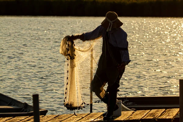 一位穿着工作服 塑料靴和水桶帽的渔夫正在准备撒网 然后在日落时把网扔进切萨皮克湾 他在一个木制的码头上 阳光从水面反射出来 — 图库照片
