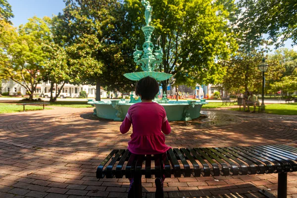 在马里兰州切斯特顿的喷泉公园 一个身穿粉色休闲装的小女孩正一个人坐在一条金属长椅上 在模糊的背景中 有一个喷泉雕像和水池 — 图库照片