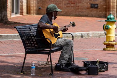Annapolis, MD 08 / 21 / 2020: Beyzbol şapkası ve maske takan yaşlı bir Afro-Amerikan adam klasik gitar çalıyor ve sokakta bir bankta otururken şarkı söylüyor. O bir sokak müzisyeni..