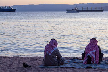 Akabe Körfezi kıyısında iki Arap adam keffiyeh giyer, agal ve thobe sahilde halı üzerinde bağdaş kurarak otururken görülürler. Kızıl denizin üzerindeki güzel günbatımına bakıyorlar..