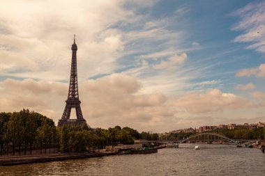 Baharda Paris, Fransa 'nın manzarası bir tarafta Seine nehri diğer tarafta da ünlü Eyfel kulesi. Arka planda küçük metal bir köprü, ağaçlar, bir nehir teknesi, binalar görülüyor..