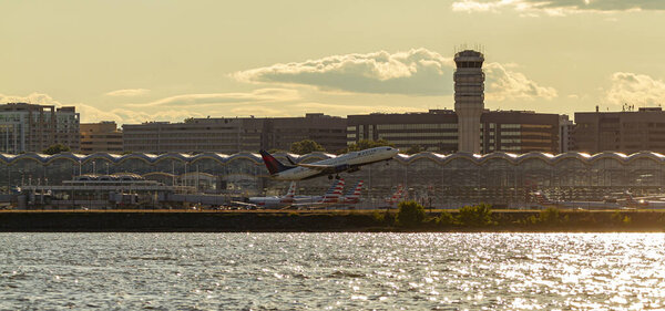 Washington DC, USA 10 / 03 / 2020: Panoramic view of Ronald Reagan National Airport across Potomac River at sunset. изображение показывает башню, терминалы, припаркованные самолеты и самолет Delta Airline, взлетающий.