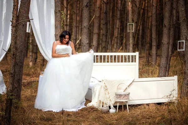 Woman is trying on wedding dress next to bed in the morning on the nature background of forest, morning of the bride concept