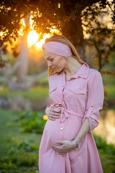 Porträtt av en lycklig och stolt gravid ung kvinna som tittar på hennes mage i parken vid soluppgången med en varm bakljus i bakgrunden, New Life Concept — Stockfoto