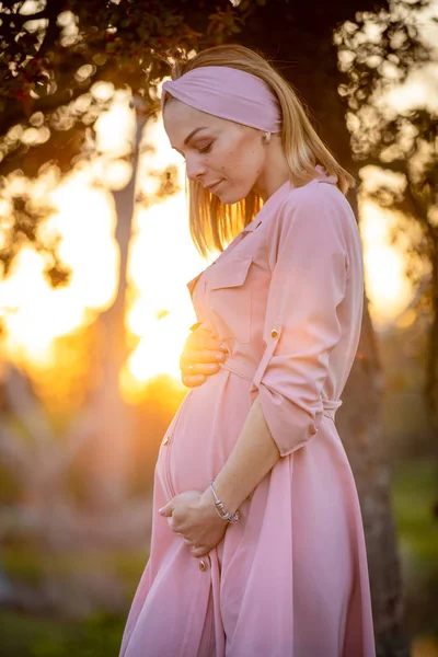 Porträtt av en lycklig och stolt gravid ung kvinna som tittar på hennes mage i parken vid soluppgången med en varm bakljus i bakgrunden, New Life Concept — Stockfoto